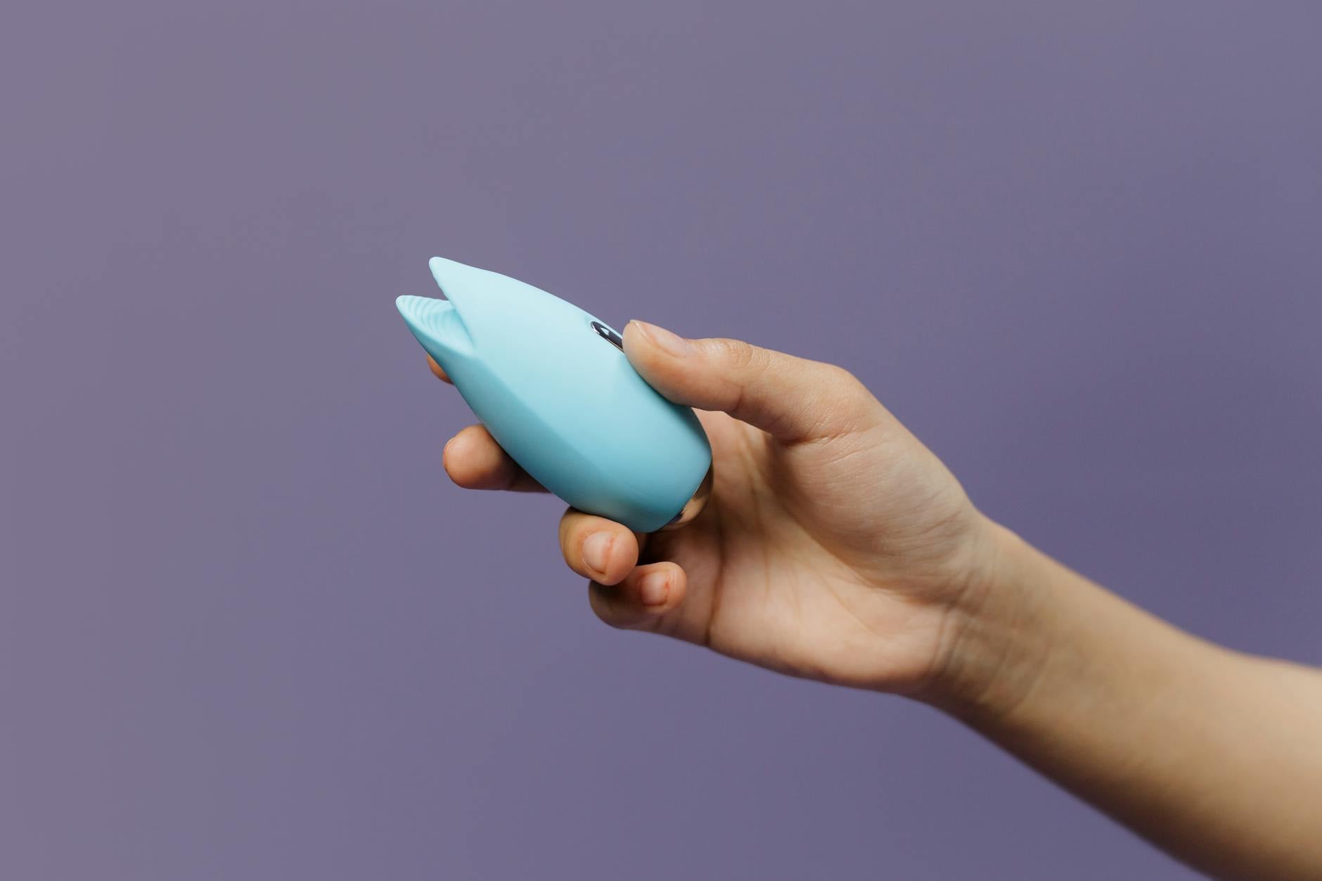 Blue silicone clitoral vibrator held in hand against purple background